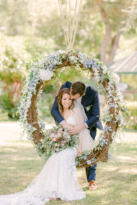 a bride sitting in a floral swing with her groom leaning over her to kiss her temple at their whimsical micro wedding at lairmont manor