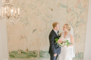 a bride and groom at lakewold gardens standing inside below a chandelier and a beautiful old mural on the wall