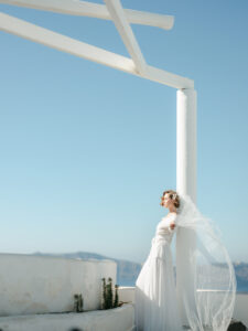 a bride standing against a white pillar holding her wedding veil as it blows in the wind