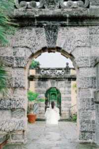 a woman in a white dress running away from the camera at vizcaya museum under some stone arches