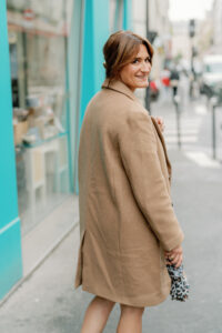 behind the lens a woman in a brown coat walking away from the camera