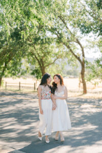 two women in white dresses standing on a road under trees at St Francis Winery in Sonoma for their engagement session
