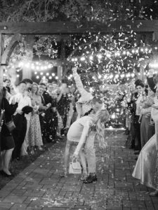  a black and white photo of a man dipping a woman in a white dress with confetti all around them for their wedding send off