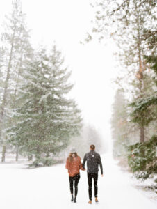 a couple holding hands walking away from the camera while its snowing with green trees all around