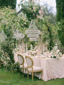 a elegant and luxurious wedding table with pink and gold accents and floral arches over the table with crystal chandeliers over the table on the lawn of villa cetinale in tuscany