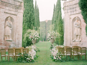 a wedding ceremony setting in between two large statues on the lawn of villa cetinale in tuscany italy with large floral arrangements and wooden chairs in rows