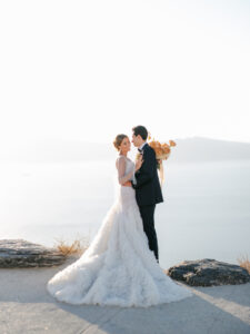 a bride and groom standing on the cliffs looking over santorini during sunset at their santorini elopement 