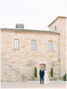 a bride and groom holding hands and walking towards the camera with sunstone winery in the background