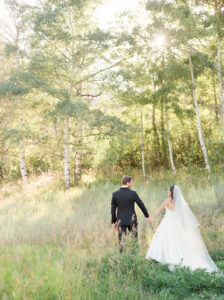 a bride and groom in a large tall grassy field with aspen trees behind them with the sun shining through