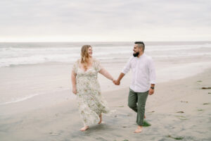 a man and woman holding hands on a beach walking at their Torrey Pines beach engagement session