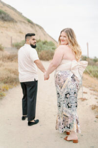 a man and woman walking away from the camera and looking back at the camera smiling with Torrey Pines beach behind them