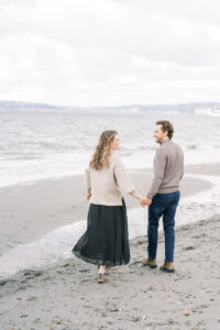 a man and woman walking on the sand at alki beach in seattle for their engagement session photoshoot