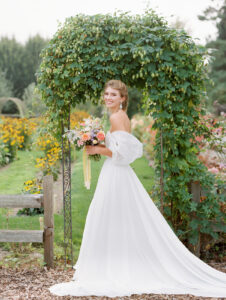 a bride holding a colorful wedding bouquet and laughing at the camera in front of a green arch at a barn wedding venue in snohomish