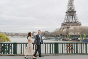 a couple walking on a bridge in paris with the seine river and the eiffel tower behind them while being photographed for their engagement photos
