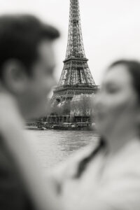 a black and white photo of a couple looking at each other with the eiffel tower in focus behind them for their engagement session in paris