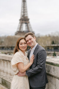 a couple looking and smiling at the camera with the eiffel tower behind them during their engagement session in paris