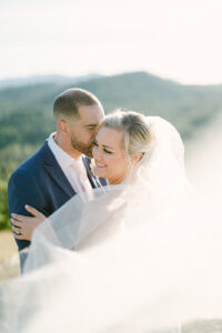a groom kissing his bride while she looks away and smiles and her veil is blowing in the wind at their wedding at swiftwater cellars