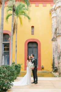 a bride and groom standing in front of a bright yellow and red building at hacienda sac chic in mexico for their destination wedding