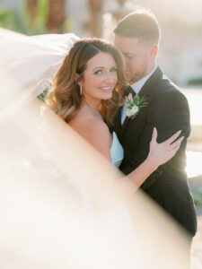 a bride and groom standing together while the wedding veil is blowing in the wind and the bride is looking at the camera and smiling