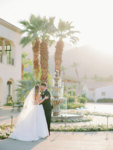 a bride and groom standing in front of a water fountain and palm trees kissing during sunset in palm springs