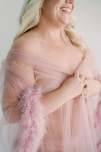 a blond woman smiling and looking away while holding a sheer pink robe with fuzzy pink sleeves for her boudoir session in seattle washington