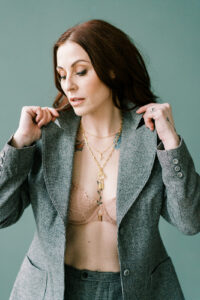 a woman wearing a blazer and a nude lace bra under it is looking at the floor and her hands are popping the collar of the blazer she's wearing
