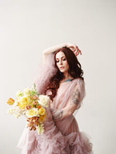 a woman wearing a soft pink fluffy see-through robe holding flowers in one hand and in another hand is resting on her head while she looks down at the bouquet