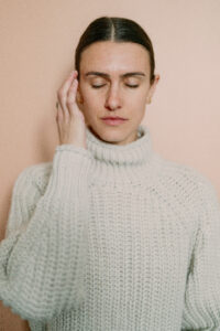 a woman in a tan sweater with her eyes closes and one hand tapping on the side of her head