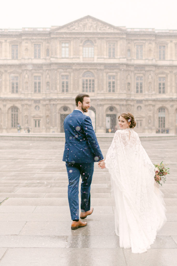 bride and groom holding hands and walking away from the camera as the bride looks back and smiles in paris as it snows