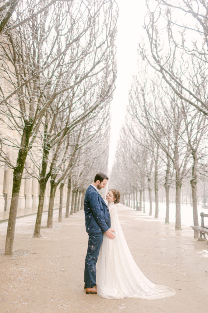 bride and groom standing facing one another in an alley of trees in Paris as it snows around them