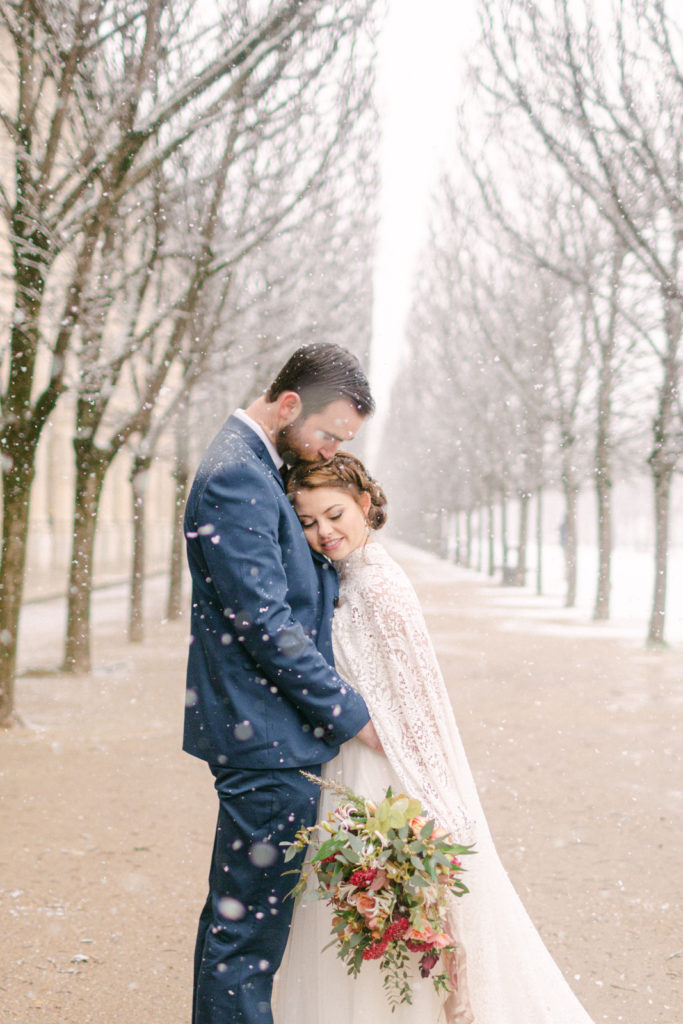 bride resting her head on her grooms chest and looking down at her flowers in Paris as it snows