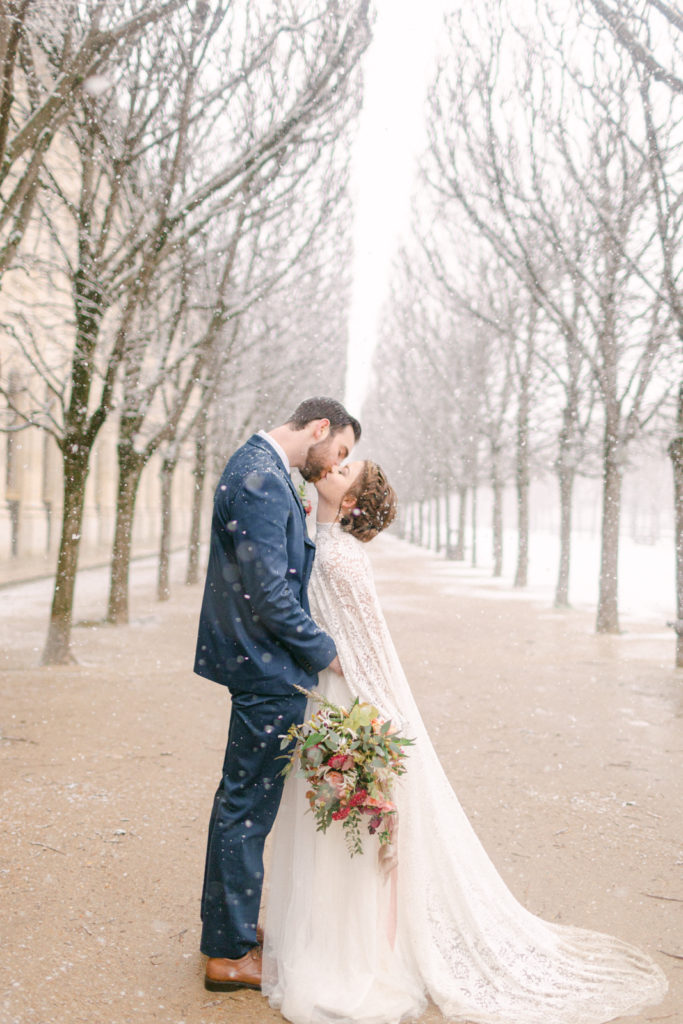 bride and groom kissing in an alley of trees in paris as it snows around them
