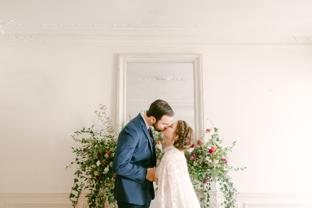Bride and groom kiss in front of a fireplace mantel with a floral arrangement in a paris apartment 