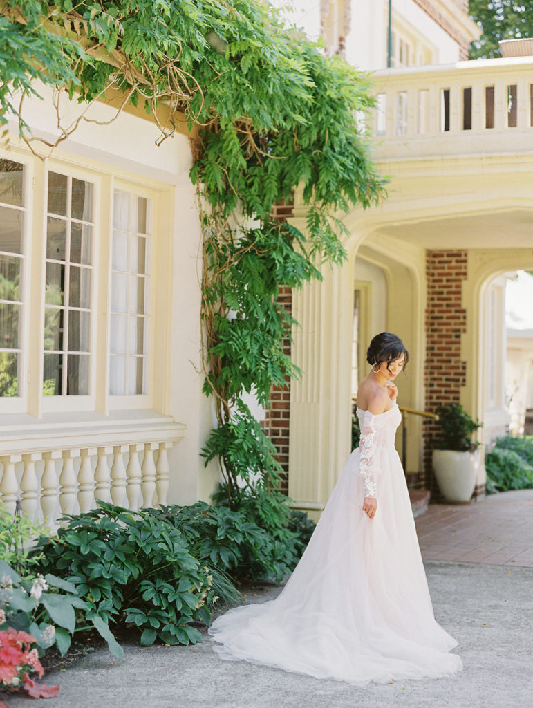 bride in front of a large window with ivy growing up it as she looks down at the ground in her pink wedding dress