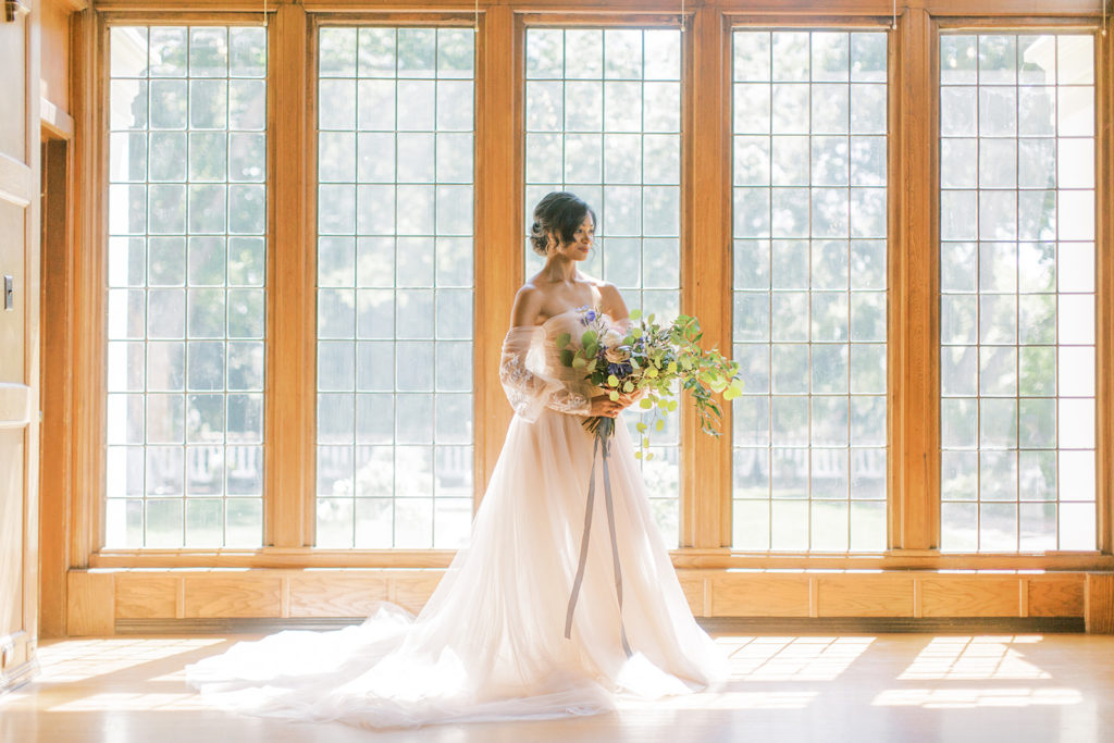 bride in a pink wedding dress holding her flowers in front of large windows