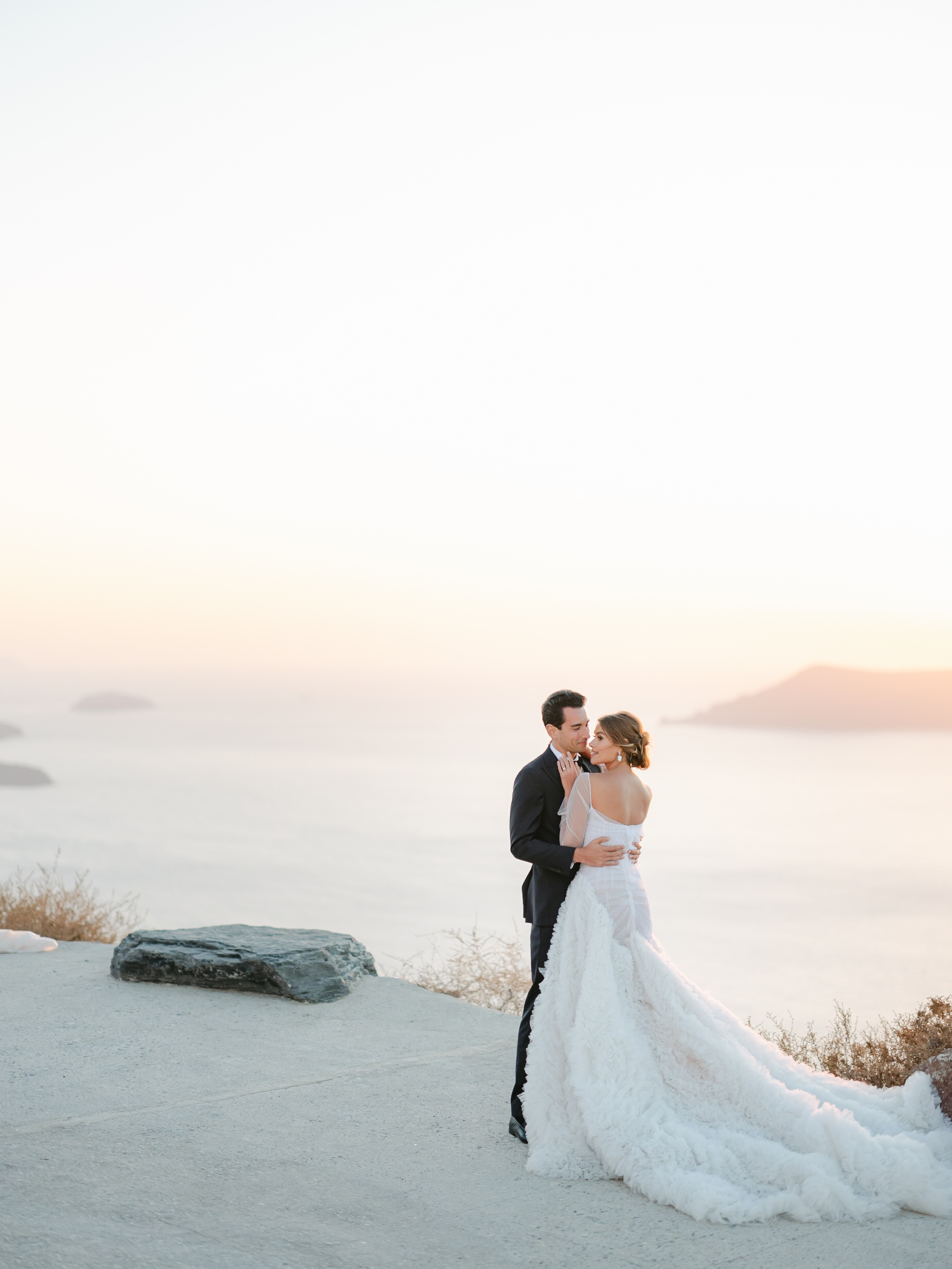 a bride and groom facing each other at their destination wedding in santorini with the ocean and sunset behind them