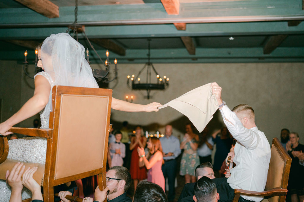 bride and groom on chairs during the horah dance at their wedding reception holding a cloth