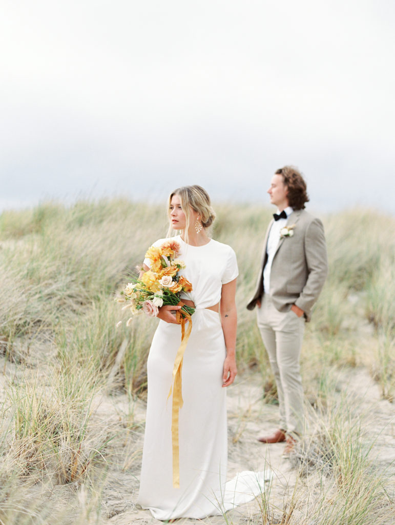 bride standing in front of groom while holding her wedding bouquet and looking off into the distance at cannon beach