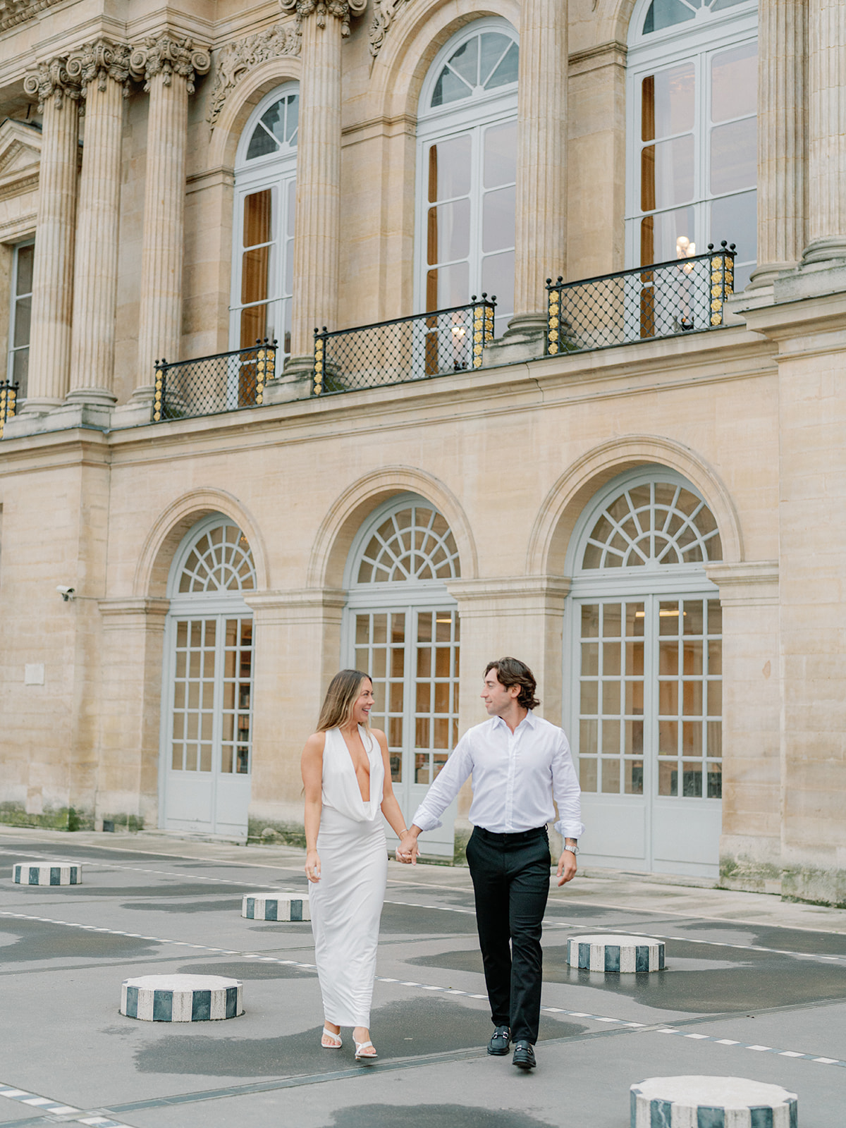 a woman in a white dress and a man in a white dress shirt and black pants walking and holding hands with black and white pillars at the grand palais in paris france