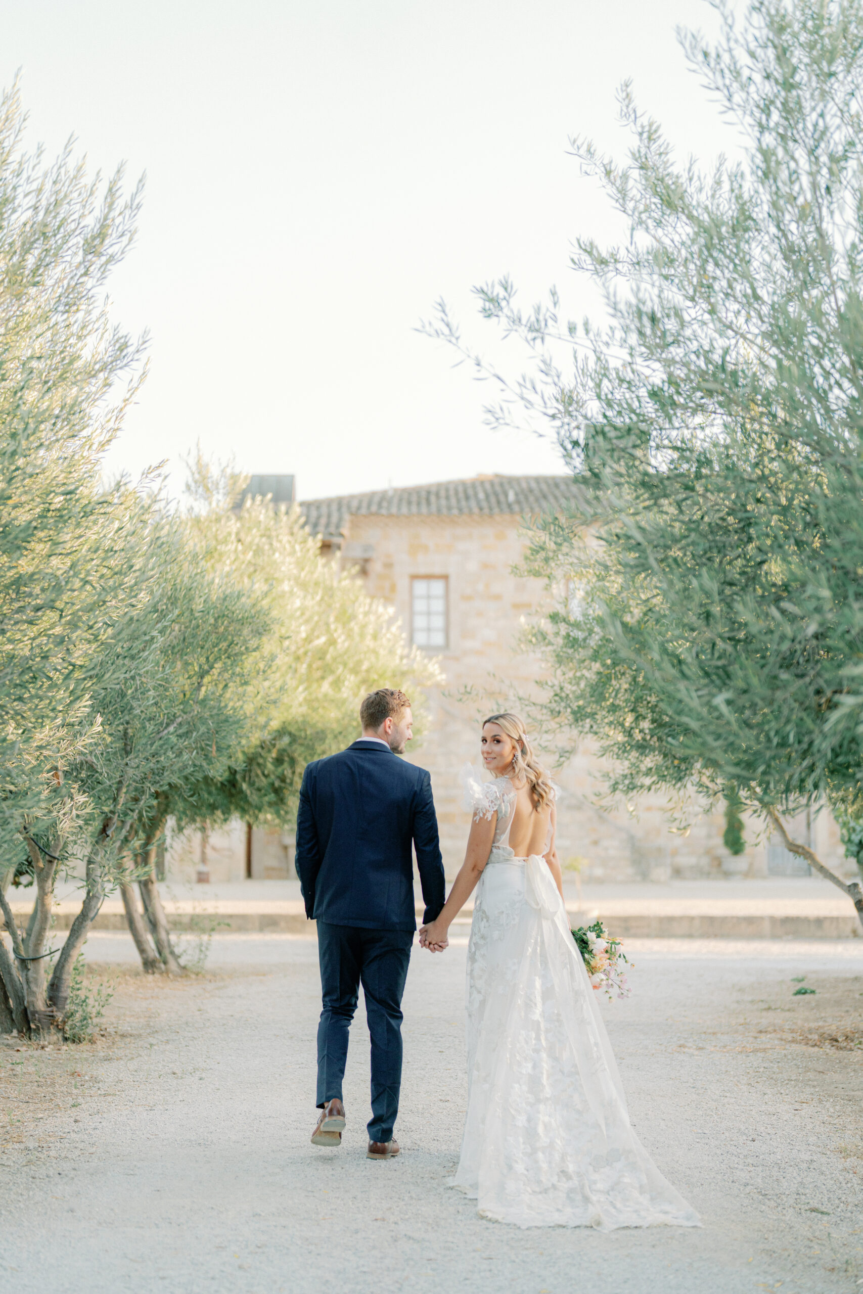 a bride and groom holding hands and walking away from the camera in a row of trees at a wedding at sunstone winery