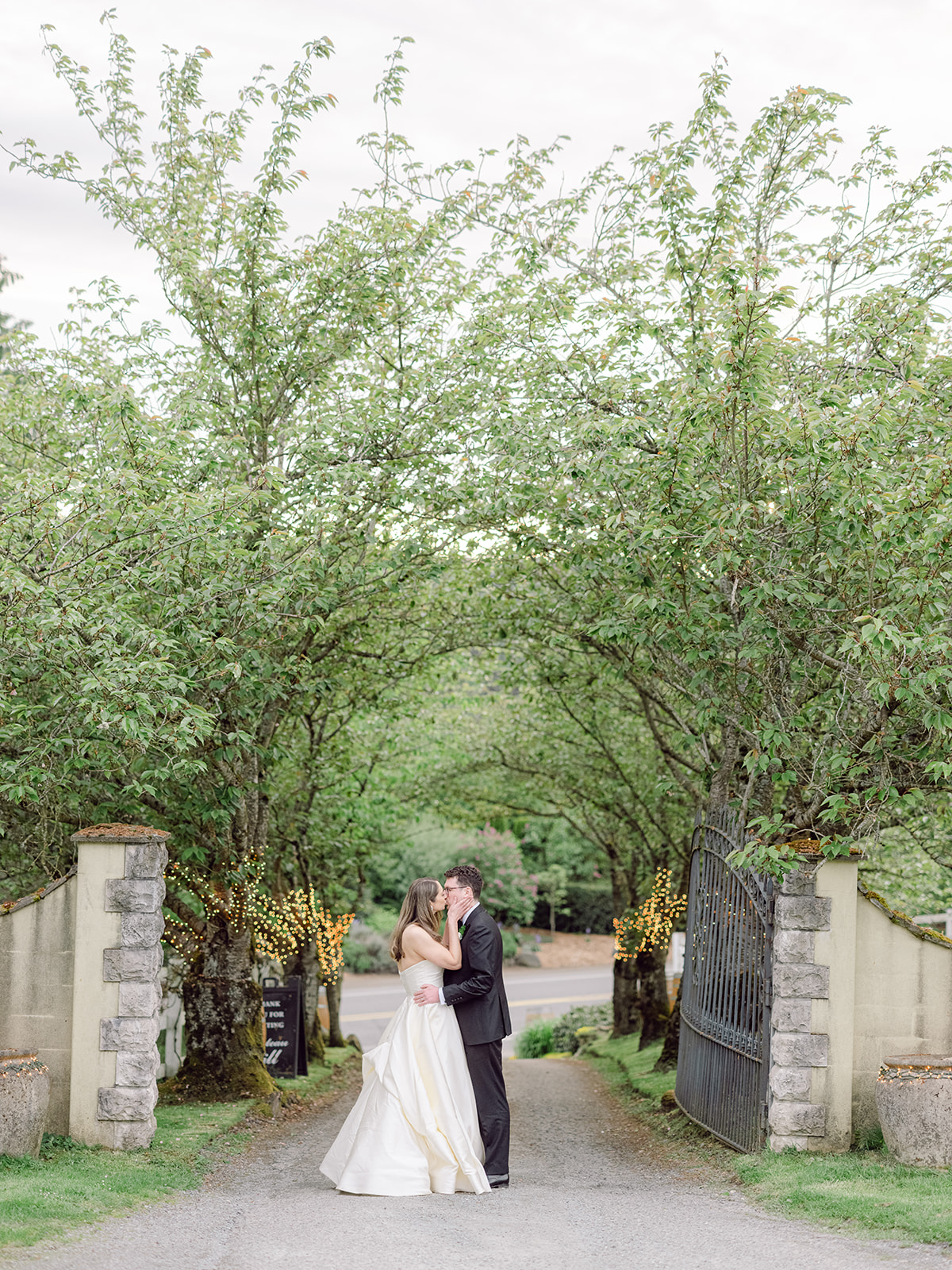a bride and groom kissing in a driveway with a row of trees behind them at their chateau lill wedding